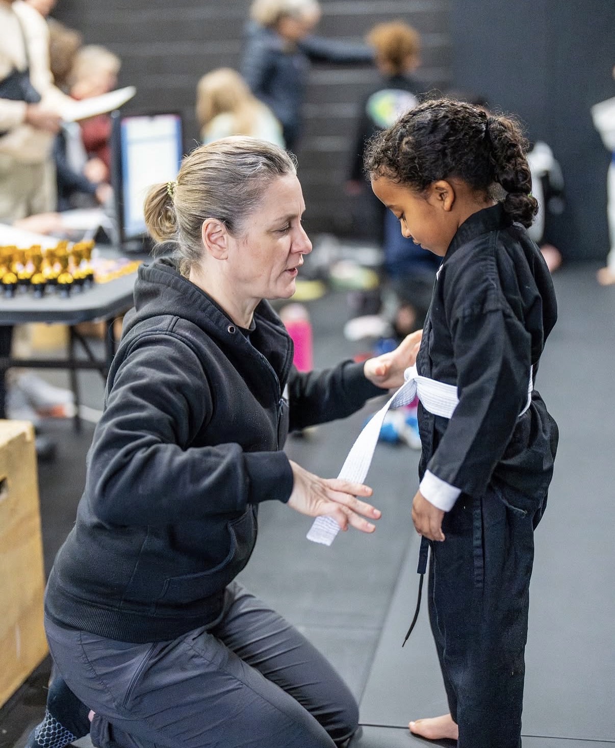Coach Kate Parish kneeling to tie a young student's white belt at WDC BJJ