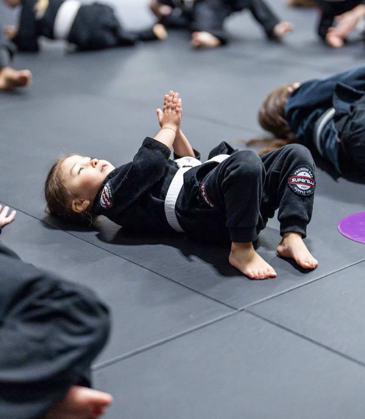 Little Grapplers toddler warm-up class — a young student in a black gi lying on the mats with hands raised during drills at WDC BJJ Takoma Park